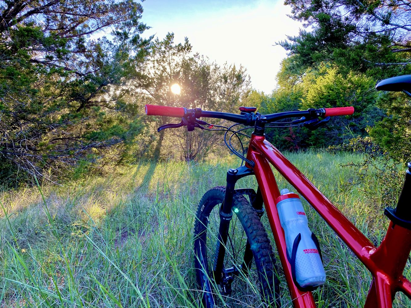 Red mountain bike sits in a grassy field looking at a setting sun through trees in the background